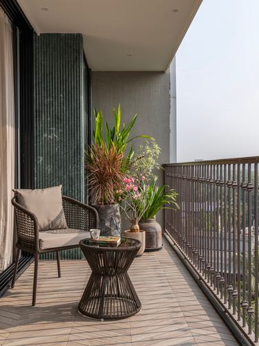 Balcony seating nook with woven chair, round table, potted plants, and slatted metal railing under soft daylight