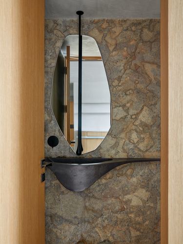 Sculptural black basin and mirror set against a mottled stone wall, framed by an oak door.