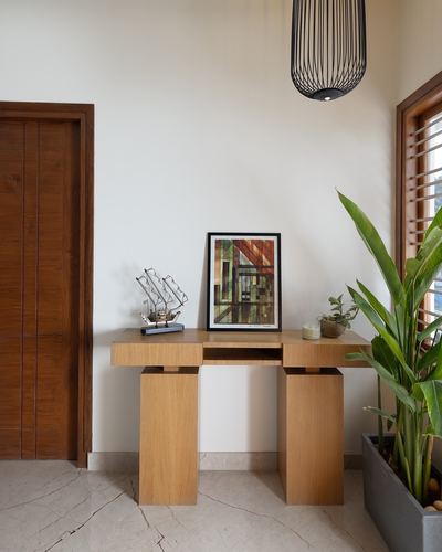 Wooden console table with framed artwork under a black wire pendant beside a wooden door and plant.