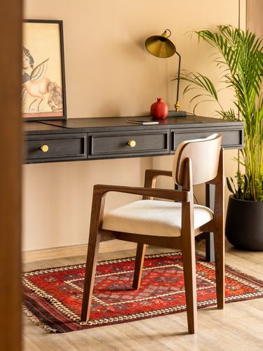 Wooden armchair with cream upholstery in front of a black console desk with brass lamp, red rug beneath.