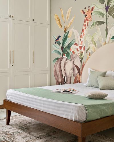 Bed with green linens and arched headboard against a jungle-themed mural beside cream wardrobes