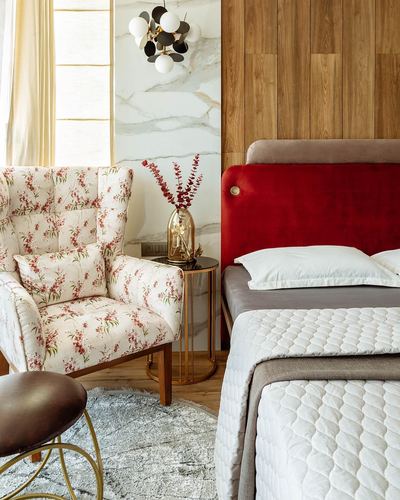 Bedroom corner with floral wingback chair, brass side table, and red velvet headboard against marble wall