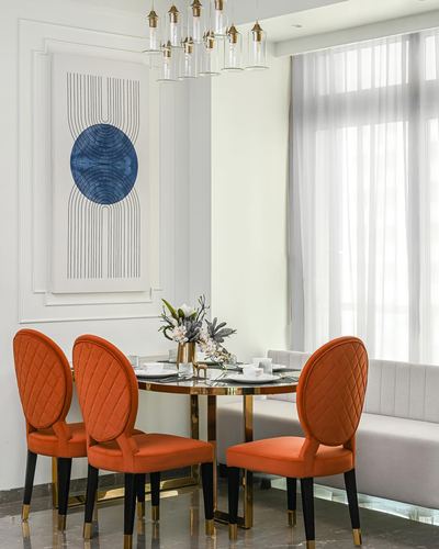 Round dining table with orange diamond-quilted chairs beneath a framed abstract print and pendant lights