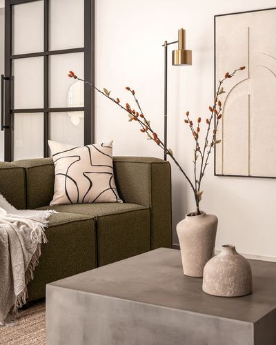 Olive boucle sofa corner with ceramic vases, branches, and concrete coffee table beside black-framed glass door