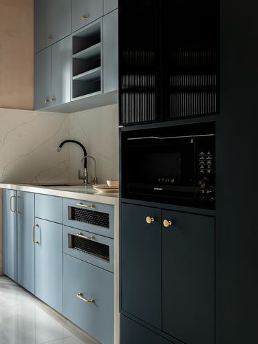 Blue and charcoal kitchen cabinetry with brass knobs and built-in microwave against marble-look backsplash