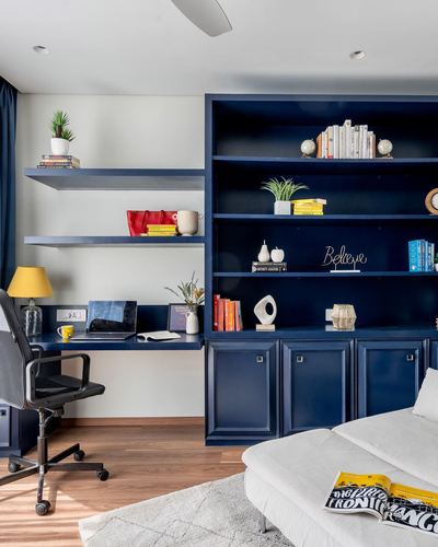 Home study with navy blue built-in shelving, floating desk, and yellow lamp beside a living room sofa