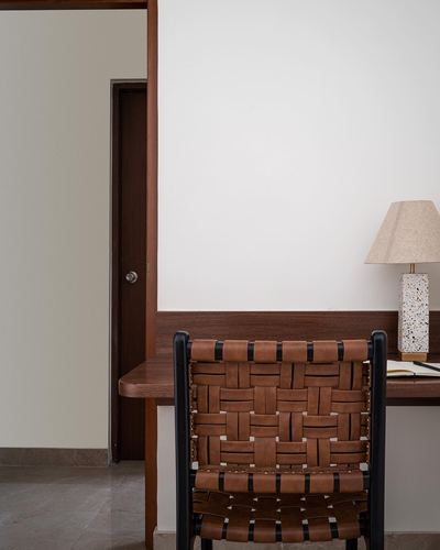 Woven leather and black metal chair at a wooden desk with terrazzo lamp, dark wood door beyond