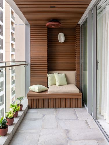 Balcony nook with slatted wood walls, built-in cushioned bench, and potted plants beside glass railing.
