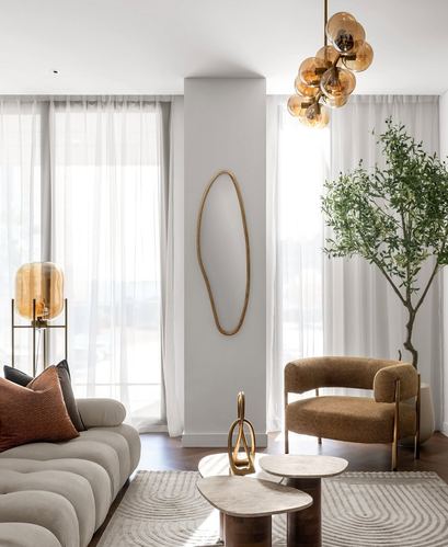 Living room with cream tufted sofa, curved tan armchair, brass mirror, amber chandelier and olive tree