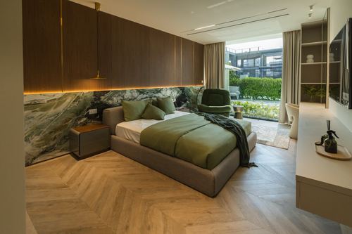 Bedroom with dark wood paneling, green stone accent wall, chevron floor and olive bedding facing a terrace window.