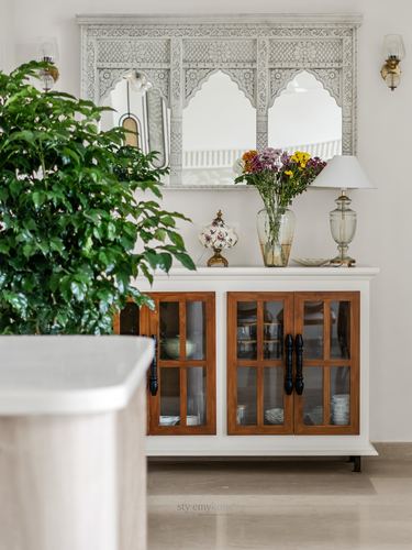 White console with wooden glass-front cabinets under a carved arched mirror, styled with flowers and a lamp