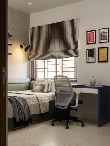 Corner bedroom with green velvet bed, striped blind, navy desk and framed posters on wall