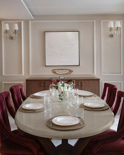 Elegant dining room with oval stone table, burgundy velvet chairs, and textured white artwork above a wooden sideboard