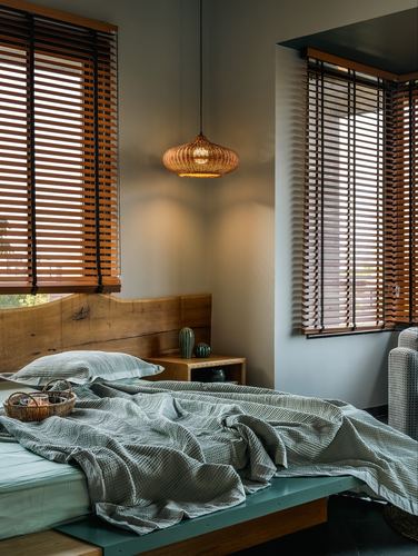 Bedroom corner with cane pendant light, wooden headboard, and rumpled sage bedding beside slatted wooden blinds