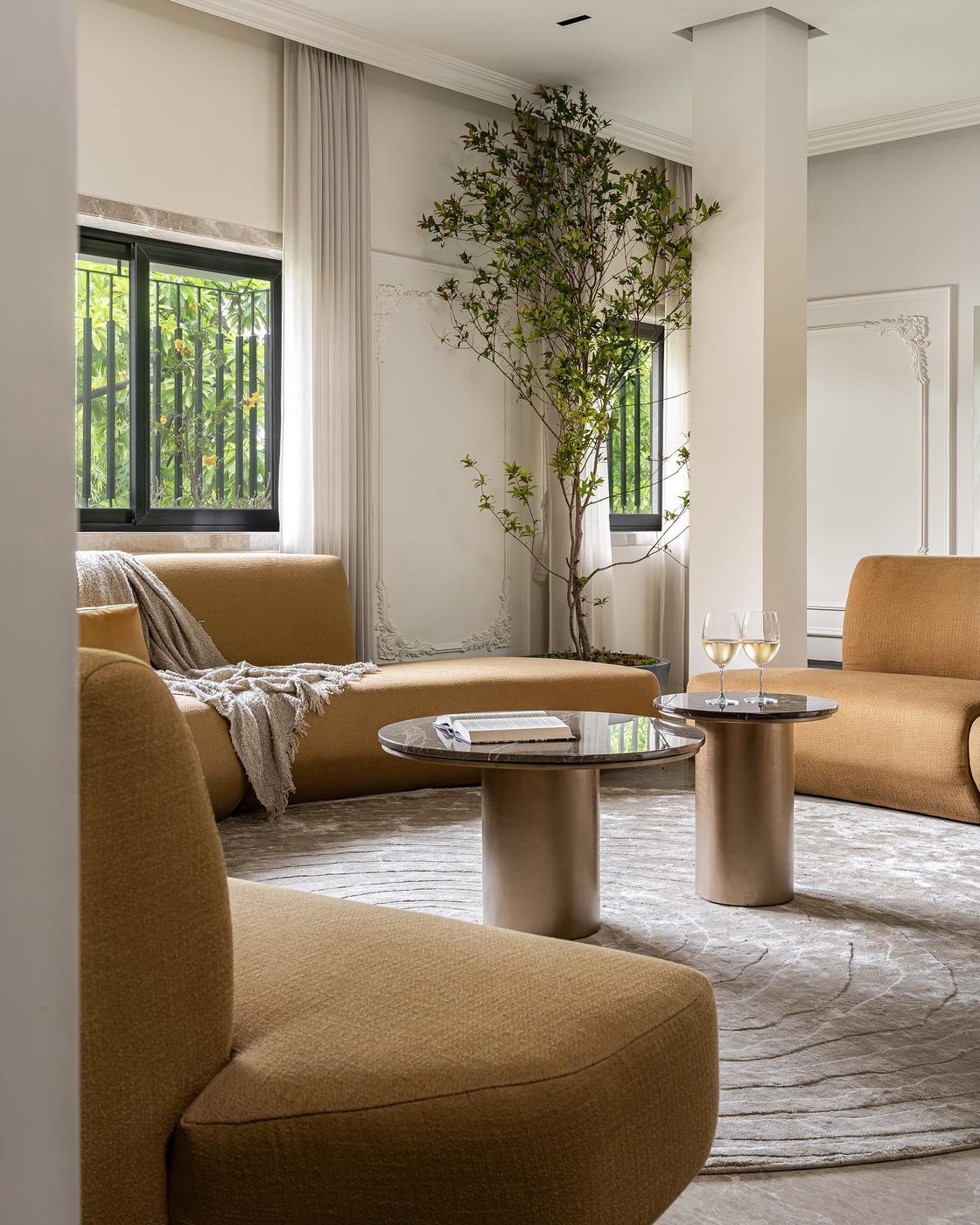 Curved mustard sofas around round stone-top tables in a neutral living room with a large indoor tree