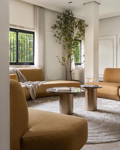Curved mustard sofas around round stone-top tables in a neutral living room with a large indoor tree