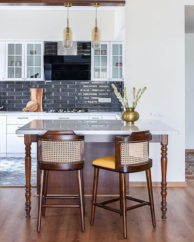 Kitchen island with cane-back barstools, dark glossy tile backsplash and brass vase under amber pendant lights