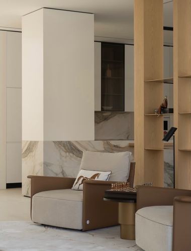 Living area with beige and leather armchairs beside a marble-clad column and oak shelving, chess table between