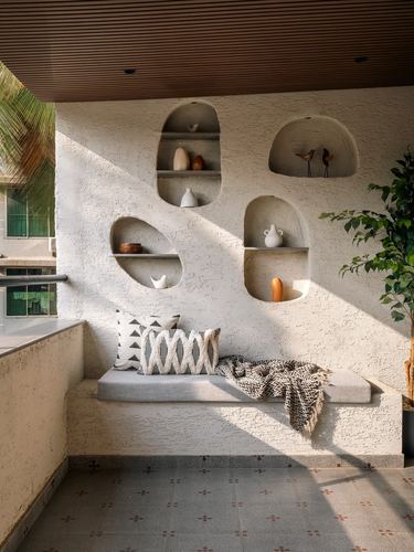Sunlit balcony with textured plaster wall, carved niches holding ceramics, and a cushioned window seat.