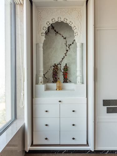 White carved arch mandir niche with marble backdrop and deity idols above built-in drawers