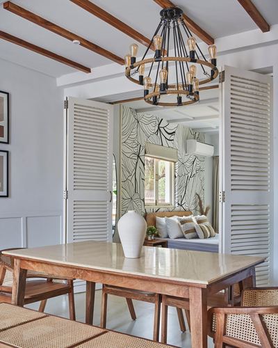 Dining room with stone-top wood table, cane chairs, and open louvered doors revealing a bedroom with patterned wallpaper.