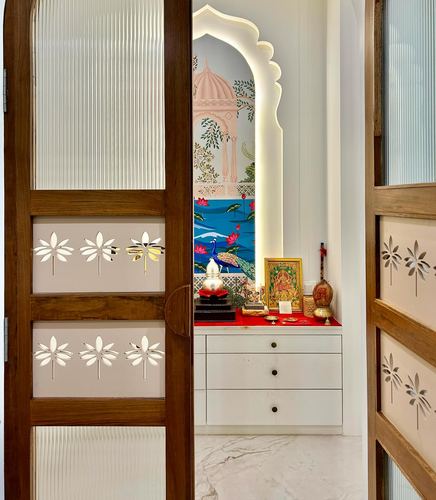 Wooden fluted door opens to reveal an arched, backlit pooja niche with mural wallpaper and brass idols