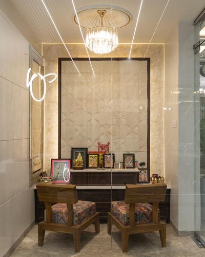 Pooja alcove with textured stone panel, crystal chandelier, deity frames, and two patterned wooden chairs