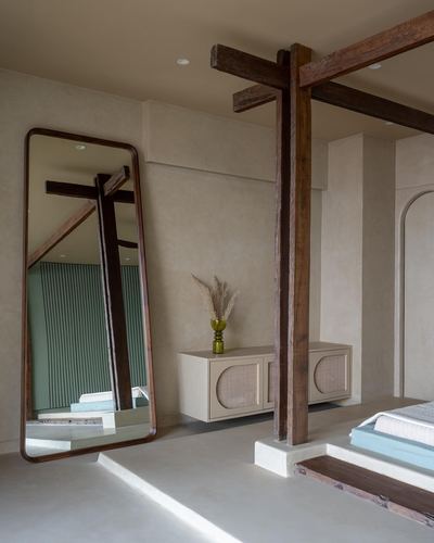 Bedroom corner with leaning wooden mirror, cane cabinet, and exposed timber beam against plaster walls