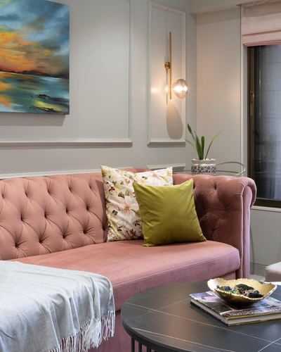 Pink tufted velvet sofa with floral and olive pillows against grey paneled wall with brass sconce