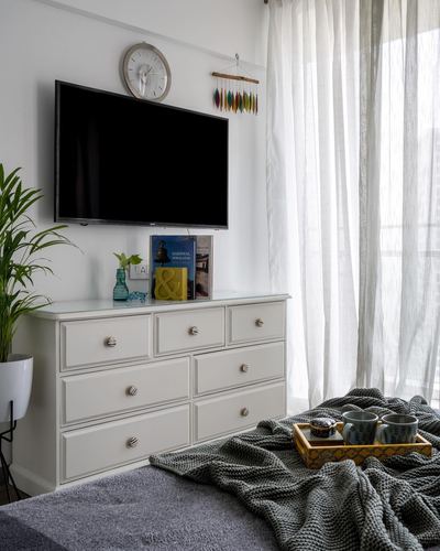 White dresser under wall-mounted tv with clock, beside sheer-curtained window in bedroom