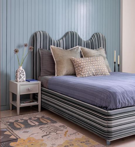 Bed with scalloped striped upholstered headboard against blue wood-paneled wall, styled with cushions and nightstand