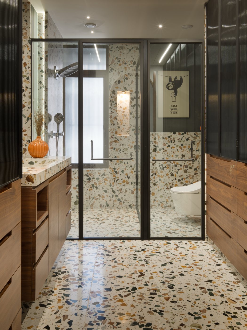 Bathroom with terrazzo floor and walls, glass shower partition, walnut vanity, and wall-mounted toilet beyond