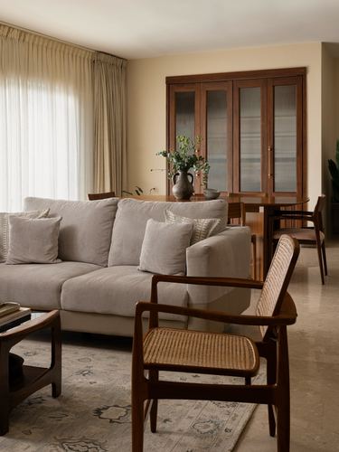 Living room with beige sofa, cane armchair, wooden cabinet, and dining table beyond, lit by sheer curtains