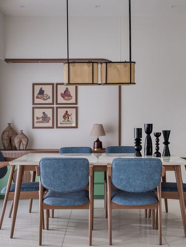Dining table with blue upholstered chairs under a cane pendant light, framed art on wall behind