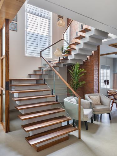 Floating wood and concrete staircase with glass rail beside exposed brick wall and armchairs