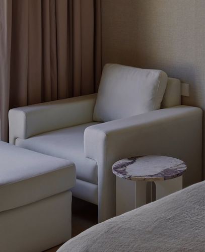 Cream armchair with cushion beside a marble-topped side table and beige curtain