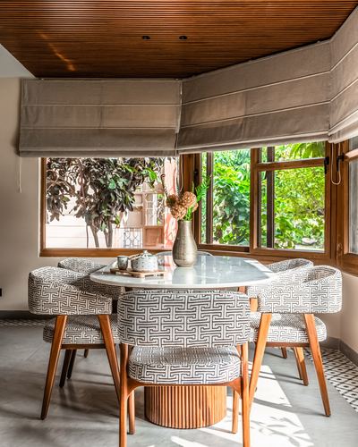 Round marble dining table with patterned chairs beside corner windows overlooking greenery, wood-slat ceiling above