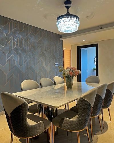 Marble dining table with quilted grey chairs under a crystal chandelier beside a chevron gold-lined accent wall
