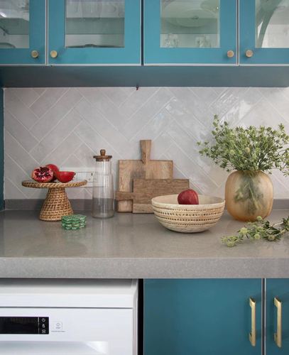 Teal kitchen counter styled with wooden boards, ceramic bowl, glass jar and vase against herringbone tile backsplash