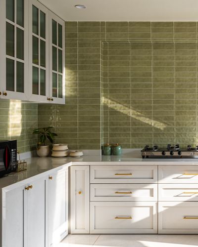 White shaker kitchen with olive green tiled backsplash and gold hardware, sunlight streaking across counter