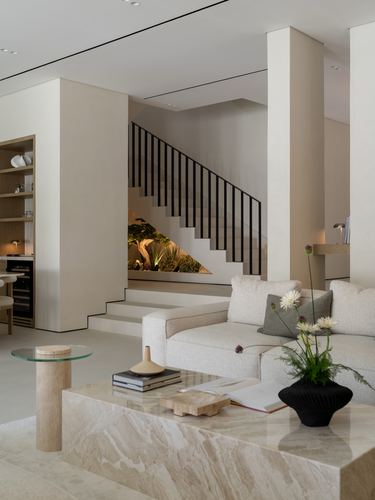 Neutral-toned living room with boucle sofa, travertine coffee table, and staircase over a lit indoor garden.