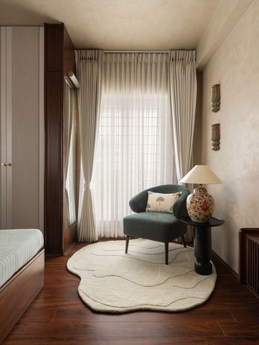 Bedroom corner with green armchair, floral lamp, and curved cream rug against sheer curtained window.