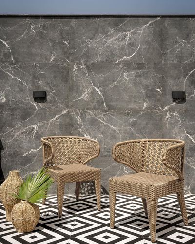 Two woven rattan chairs on black and white geometric tile against a grey marble wall outdoors