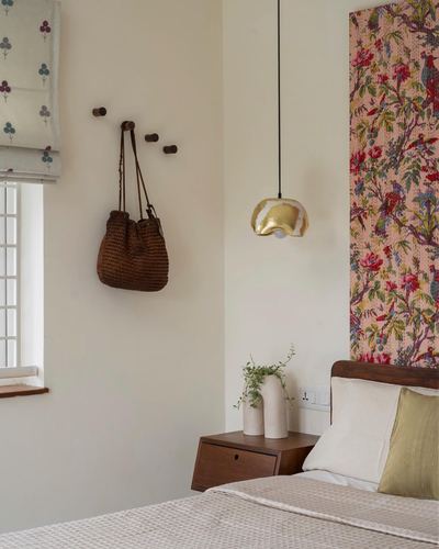 Bedroom corner with woven bag on wooden pegs, gold pendant light, and floral fabric headboard panel