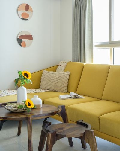 Mustard yellow corner sofa with cushions, round wooden coffee table, sunflowers, and circular abstract wall art.