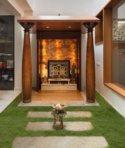 Pooja alcove with carved wooden columns, backlit amber stone panel, and a grass courtyard path leading to it