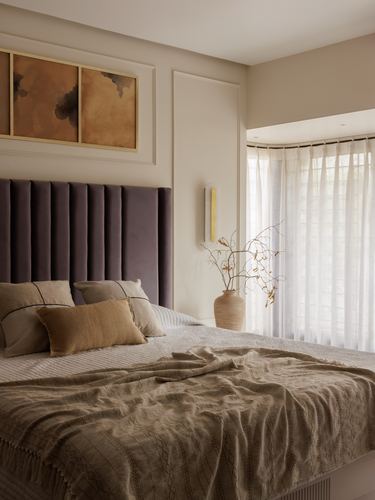 Bedroom with fluted mauve headboard, layered neutral linens, and sheer-curtained bay window with soft daylight