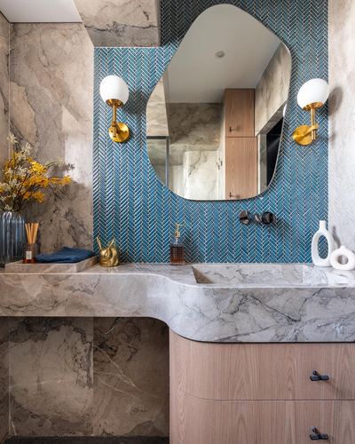 Bathroom vanity with curved marble counter, teal herringbone tile, organic mirror, and brass sconces