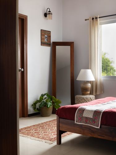 Bedroom corner with wooden mirror, cane lamp base, plant, and red-draped bed near window
