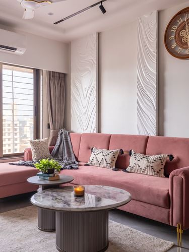 Pink velvet sectional sofa with patterned cushions beneath textured wall panels and round marble coffee tables